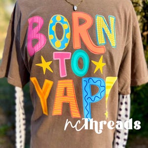 May include: A brown t-shirt featuring the phrase "BORN TO YAP" in vibrant, bubble-style lettering. The shirt has a layered design, with a white sleeve patterned with black polka dots visible. The brand name "ncthreads" is printed at the bottom.