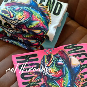 May include: Stacked t-shirts featuring a vibrant, cartoon-style fish wearing sunglasses and a hat. The shirts are white, teal, and pink, with the words "WEEKEND" and "HOOKED" in bold letters. The fish design is colorful with shades of blue, green, yellow, and red.