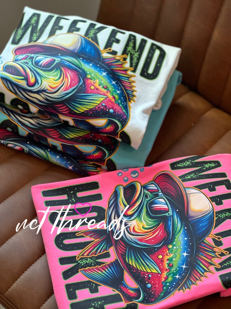 May include: Stacked t-shirts featuring a vibrant, cartoon-style fish wearing sunglasses and a hat. The shirts are white, teal, and pink, with the words "WEEKEND" and "HOOKED" in bold letters. The fish design is colorful with shades of blue, green, yellow, and red.