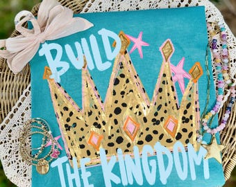 May include: A square teal sign featuring a gold crown with a black leopard print pattern, pink star accents, and the white text "BUILD THE KINGDOM". A light beige ribbon bow is attached to the top left corner, and beaded jewelry rests on the sign.