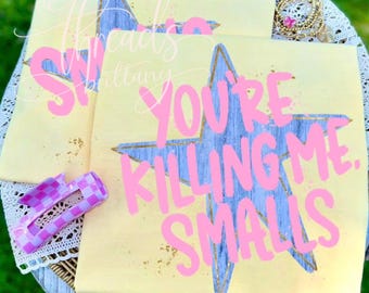 Puede incluir: Una camiseta de color amarillo pálido con una gran estrella azul con textura de veta de madera y contornos dorados. El texto en rosa brillante dice "YOU'RE KILLING ME, SMALLS". Una pinza para el pelo a cuadros morados y blancos está colocada a la izquierda.