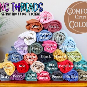 Puede incluir: Una pila de camisetas enrolladas en varios colores, incluyendo azul, verde, rojo, amarillo y rosa. Las camisetas est&aacute;n etiquetadas con nombres de colores, como "Seafoam", "Berry" y "Mustard". El texto "COMFORT C1717 COLORS" es visible en la parte superior de la imagen.