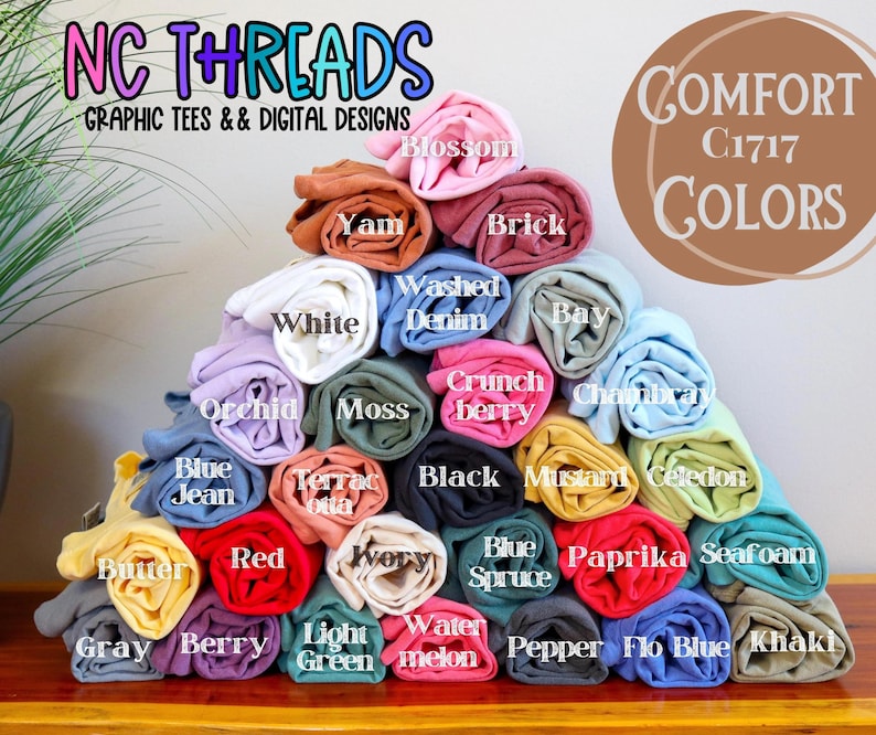 Puede incluir: Una pila de camisetas enrolladas en varios colores, incluyendo azul, verde, rojo, amarillo y rosa. Las camisetas est&aacute;n etiquetadas con nombres de colores, como "Seafoam", "Berry" y "Mustard". El texto "COMFORT C1717 COLORS" es visible en la parte superior de la imagen.