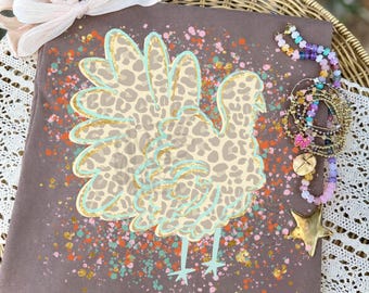 May include: A brown t-shirt featuring a turkey graphic with a tan leopard print pattern, outlined in light blue. The design includes orange, pink, and gold paint splatters. A light pink ribbon bow is attached to the top, and a multi-colored beaded bracelet with a gold star charm rests on the right side.