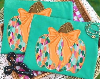 May include: Two teal fabric pouches featuring a graphic of a pumpkin with a large orange bow. The pumpkin has a multicolored pattern of pink, orange, teal, and white spots. A black hair accessory with white polka dots is placed in the corner.
