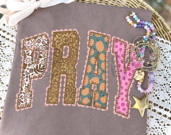 May include: A brown t-shirt featuring the word PRAY in large, appliqued letters. Each letter has a distinct pattern: floral, leopard print, orange pumpkins on teal, and pink polka dots. The letters are outlined with pink lace trim.