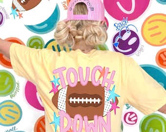 Puede incluir: Una camiseta de color amarillo pálido con un gráfico de balón de fútbol americano marrón con extremos de lunares blancos, rodeado de estrellas rosas y azules y el texto "TOUCH DOWN" en letras rosas. Se lleva una gorra de béisbol a rayas.