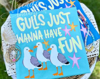 Puede incluir: Una camiseta azul brillante con tres gaviotas blancas de alas grises y picos naranjas. El texto GULLS JUST WANNA HAVE FUN está impreso en verde claro y blanco. El diseño incluye estrellas de mar rosas y naranjas sobre la tela.