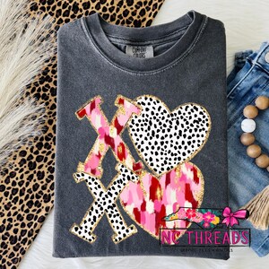 Puede incluir: Camiseta Comfort Colors gris oscuro con un diseño de San Valentín. El diseño presenta las letras XOXO en estampado de leopardo rosa y negro, y dos corazones con pinceladas rosas y rojas y lunares negros. El diseño está delineado con purpurina dorada.