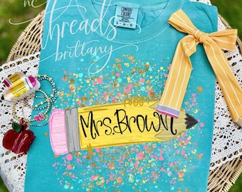 Puede incluir: Una camiseta de manga corta color verde azulado con un gráfico de un lápiz amarillo con goma de borrar rosa y el texto Mrs. Brown. El diseño está rodeado de salpicaduras de pintura dorada y rosa. Un lazo a rayas amarillas y blancas está atado al hombro derecho.