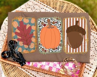 May include: A brown t-shirt featuring three rectangular graphic panels. The left panel shows a red leaf over a blue pumpkin pattern. The center panel displays an orange pumpkin over a floral print. The right panel depicts a brown acorn over stripes.