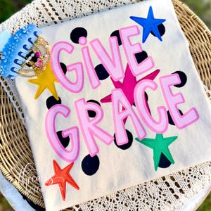 May include: Cream-colored t-shirt with the words "GIVE GRACE" in pink lettering, accented with black polka dots and colorful star shapes. A blue hair clip and jewelry are visible in the upper left corner.