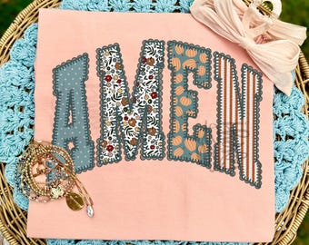 May include: A peach-colored t-shirt featuring the word AMEN in large, block-style letters. Each letter has a distinct pattern: polka dots, floral print, pumpkin print, and vertical stripes. A stack of gold-toned bracelets and a light pink hair bow.