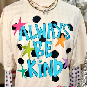Puede incluir: Camiseta color crema con las palabras "ALWAYS BE KIND" en turquesa, acentuada por estrellas coloridas y lunares negros. La camiseta tiene cuello redondo y mangas cortas. El logo de NC Threads está en la parte inferior.