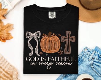 May include: A black t-shirt featuring a graphic print with a white line-art bow, an orange pumpkin, and a white line-art cross. The text reads GOD IS FAITHFUL in every season. The shirt has a Comfort Colors label indicating a size large.