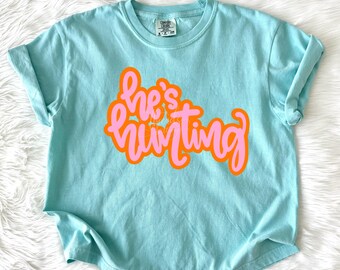 May include: A light blue short-sleeve t-shirt with a crew neckline. The center of the shirt features the text "he's hunting" in a stylized, cursive font with pink lettering outlined in a bold orange color. The shirt has a size small tag.