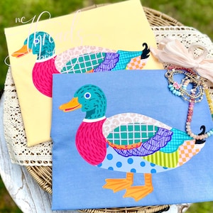 May include: Two t-shirts with duck designs. One is yellow, the other is blue. Each shirt features a colorful duck illustration with a teal head, pink chest, and patterned wings. The ducks have orange beaks and feet. The text "nc" and "Aprils" is visible.