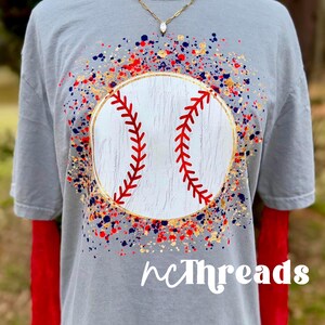 May include: A gray t-shirt featuring a baseball design. The baseball is white with red stitching and a gold outline, surrounded by a splatter design in red, blue, and gold. The text "nCThreads" is at the bottom.