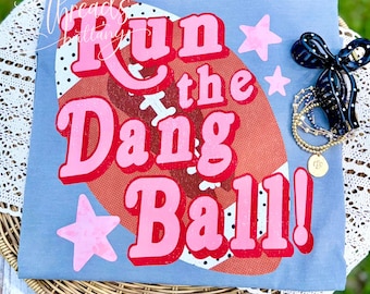 Puede incluir: Una camiseta azul claro con un gráfico grande de un balón de fútbol americano marrón con puntos blancos y estrellas rosas. El texto "Run the Dang Ball!" está impreso en letras rojas y rosas. Un lazo negro y una pulsera dorada están al lado.