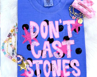May include: A periwinkle blue t-shirt featuring the text DON'T CAST STONES in large, light pink letters with a distressed texture. The design includes black polka dots and pink star shapes. A beaded necklace and a pink checkered hair clip are present.