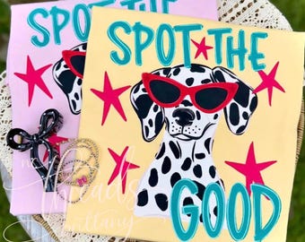 Puede incluir: Una camiseta amarilla clara con un dálmata que lleva gafas de sol rojas, el texto SPOT THE GOOD en turquesa y estrellas rosas. Una camiseta rosa clara se encuentra debajo, junto con una pinza para el pelo negra y pulseras doradas.