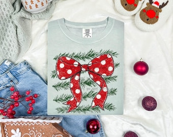 May include: A light green t-shirt featuring a large red bow with white polka dots layered over green pine branches. The shirt has a Comfort Colors tag at the neck. The design is centered on the chest, showcasing a festive holiday aesthetic.