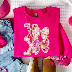 May include: A bright pink sweatshirt with the letters "XO" and a bow design. The letters and bows have a gold glitter outline and a colorful, patterned fill. The sweatshirt is displayed with blue jeans, a pink visor, and a brown shoe.