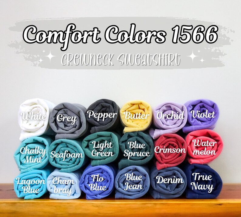 May include: A display of Comfort Colors 1566 crewneck sweatshirts in various colors, including white, grey, and blue. Each folded sweatshirt is labeled with its color name. The text "Comfort Colors 1566 Crewneck Sweatshirt" is at the top.