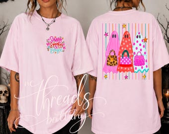 May include: A light pink t-shirt featuring a small graphic on the front chest with the text Sassy Spooky & Boujee. The back displays three ghosts in pink, red, and white with polka dots, each carrying a different handbag, set against vertical stripes.