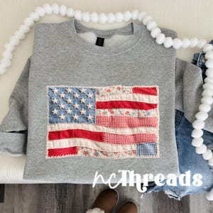 Könnte beinhalten: Ein graues Sweatshirt mit einem Patchwork-Design der amerikanischen Flagge. Die Flagge hat ein blaues Sternenfeld, rote und weiße Streifen sowie Blumen- und Vichy-Muster. Der Text "hcThreads" ist unten sichtbar.