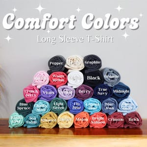 May include: A display of rolled-up Comfort Colors long sleeve t-shirts in various colors, including black, white, blue, and red. The image features the text "Comfort Colors" and "Long Sleeve T-Shirt" in white, with each shirt labeled with its color name.