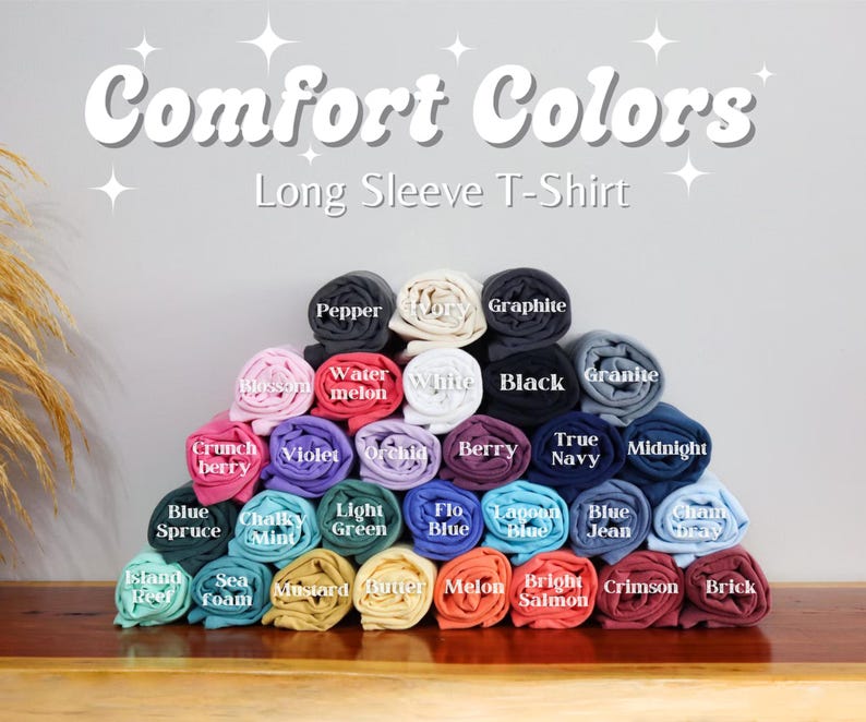 May include: A display of rolled-up Comfort Colors long sleeve t-shirts in various colors, including black, white, blue, and red. The image features the text "Comfort Colors" and "Long Sleeve T-Shirt" in white, with each shirt labeled with its color name.