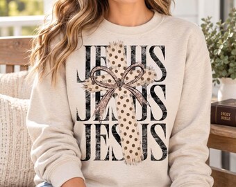 May include: A beige crewneck sweatshirt featuring a large graphic print on the front. The design includes the word JESUS repeated in black distressed lettering, overlaid with a tan polka-dot cross and a glittery brown bow tied at the center.