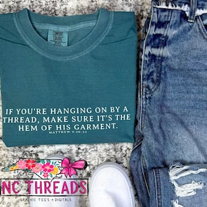 May include: Teal blue t-shirt with white text that reads "If you're hanging on by a thread, make sure it's the hem of his garment. Matthew 9:20-22". The shirt is lying on a light grey surface with a white shoe and blue jeans in the background.