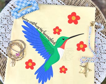 May include: A pale yellow t-shirt featuring a graphic of a green and blue hummingbird with red accents, surrounded by five red flowers with yellow centers. The text "busy watching my hummingbirds" is printed in black script above the bird.