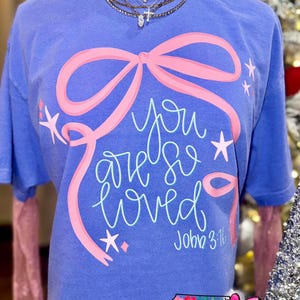 May include: A periwinkle blue t-shirt featuring a pink bow design and the words "you are so loved" in a script font. The text "John 3:16" is below the main text. The shirt has a graphic from NC Threads.
