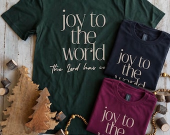 May include: A dark green, a black, and a maroon t-shirt, each featuring the text "joy to the world" in a light-colored serif font. The large green shirt also includes the additional text "the Lord has come" in a cursive script font.
