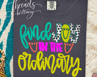 Puede incluir: Una camiseta gris carbón con el texto "find JOY IN THE ORDINARY" en turquesa, lunares blancos y negros, rosa neón y amarillo. La palabra "JOY" incluye una cara sonriente amarilla. La camiseta tiene una etiqueta de la marca Comfort Colors.