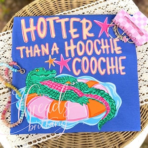 May include: Blue t-shirt with the text "HOTTER THAN A HOOCHIE COOCHIE" in pink and orange. A green alligator floats on a pink and orange inner tube. A few bracelets are visible.