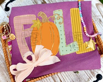 May include: A plum-colored t-shirt featuring the word FALL in large, patterned letters with an orange pumpkin replacing the letter A. A light pink ribbon bow is attached to the bottom, and a beaded necklace with a gold star charm rests on top.