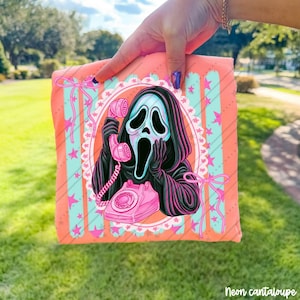 May include: A coral-colored t-shirt featuring a graphic of a black-hooded figure with a white mask holding a pink rotary telephone. The design is framed by a white lace-style border, light blue vertical stripes, pink stars, and pink ribbon bows.