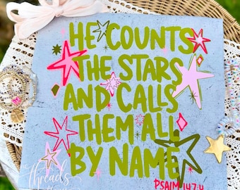 Puede incluir: Un letrero cuadrado azul claro con el texto en verde oliva HE COUNTS THE STARS AND CALLS THEM ALL BY NAME PSALM 147:4. El diseño está decorado con varias formas de estrellas rosas y verde oliva y un gran lazo rosa pálido en la esquina superior.