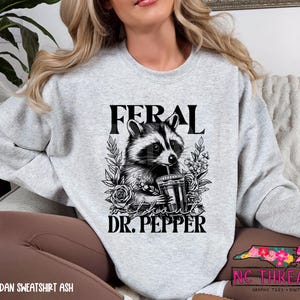 Puede incluir: Una sudadera gris con una ilustraci&oacute;n en blanco y negro de un mapache sosteniendo una lata de Dr. Pepper. El texto "Feral about Dr. Pepper" est&aacute; impreso en la sudadera.