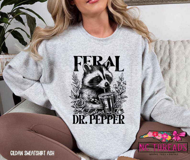 Puede incluir: Una sudadera gris con una ilustraci&oacute;n en blanco y negro de un mapache sosteniendo una lata de Dr. Pepper. El texto "Feral about Dr. Pepper" est&aacute; impreso en la sudadera.