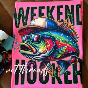 May include: A bright pink t-shirt featuring a colorful, cartoon-style fish wearing sunglasses and a backwards baseball cap. The words "WEEKEND HOOKER" are printed in large, black letters. The text "net threads" is also visible.
