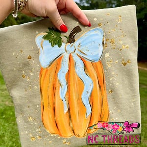 May include: A sage green t-shirt featuring a central graphic of an orange pumpkin adorned with a light blue bow and a green leaf. The design is surrounded by gold foil-like speckles. The bottom right corner displays a logo with the text NC THREADS.