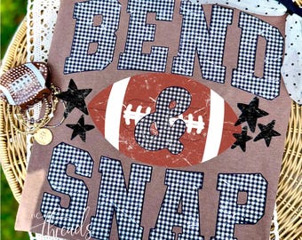 Puede incluir: Una camiseta marrón con el texto BEND & SNAP en un patrón de cuadros vichy blanco y negro. Un gráfico de balón de fútbol americano marrón con cordones blancos se sitúa en el centro, y estrellas negras están dispersas por el diseño frontal.