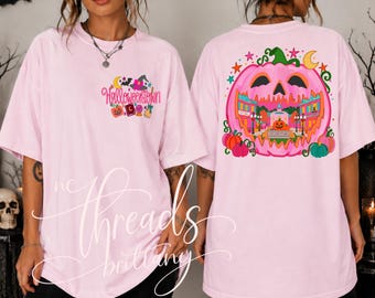 May include: A light pink short-sleeve t-shirt featuring a small graphic on the front left chest with the text Halloweentown and various icons. The back displays a large pink pumpkin graphic containing a town scene with colorful buildings.