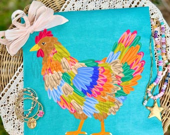 May include: A bright turquoise t-shirt featuring a colorful, stylized chicken illustration with feathers in shades of orange, yellow, blue, and green. A pale pink ribbon bow is attached to the top left, and a beaded necklace with a gold star charm.