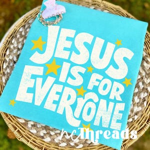 May include: A light blue t-shirt with the text "JESUS IS FOR EVERYONE" in white, accented with yellow stars. The shirt is displayed on a woven basket. A hair clip and bracelet are on top of the shirt.